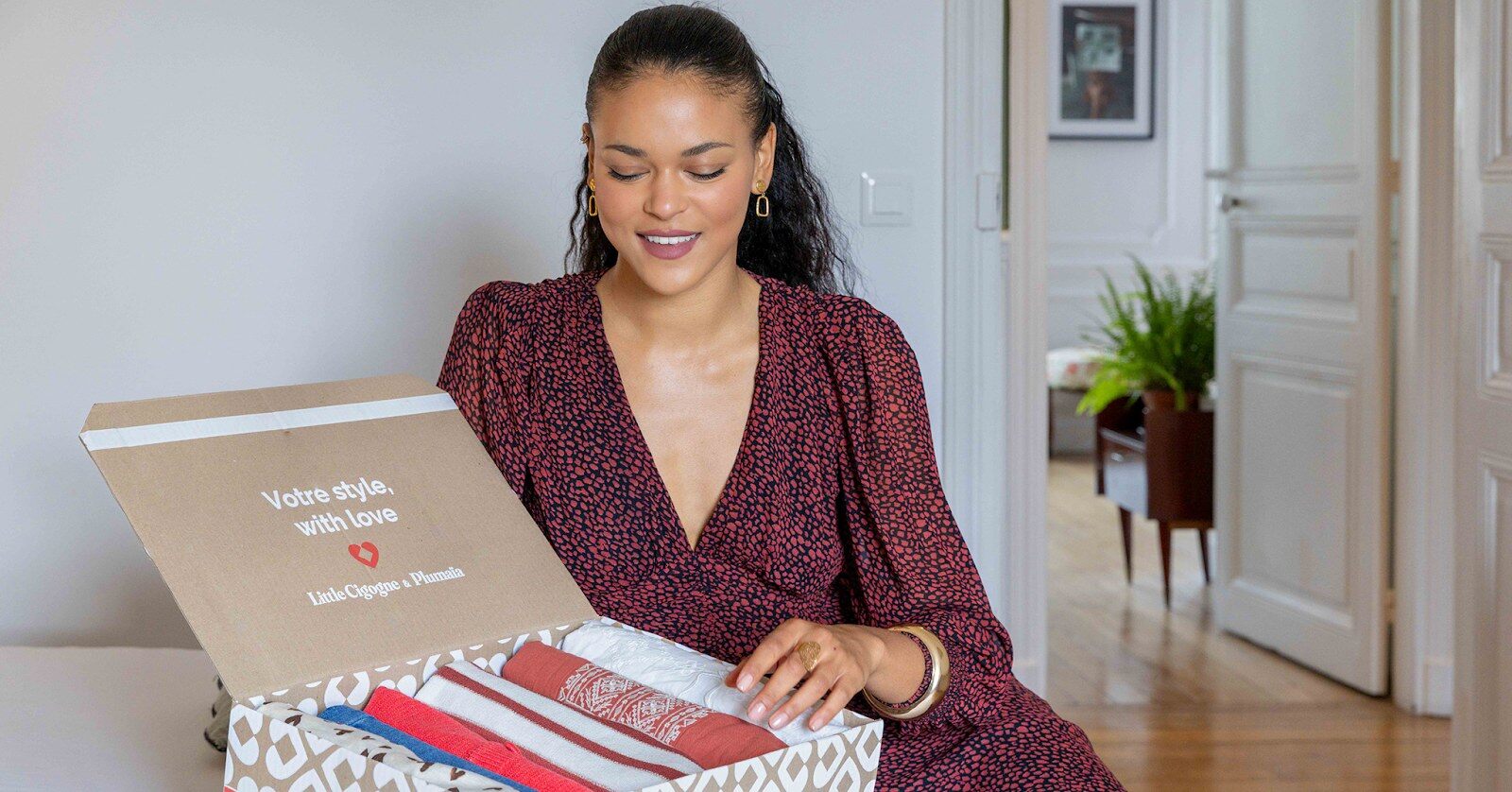 Une femme souriante ouvre une box de vêtements Little Cigogne x Plumaïa dans son salon