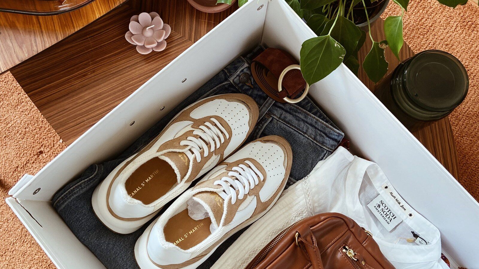 Une box La Malle Française ouverte avec des baskets blanches, un jean, une chemise blanche et un sac en cuir camel