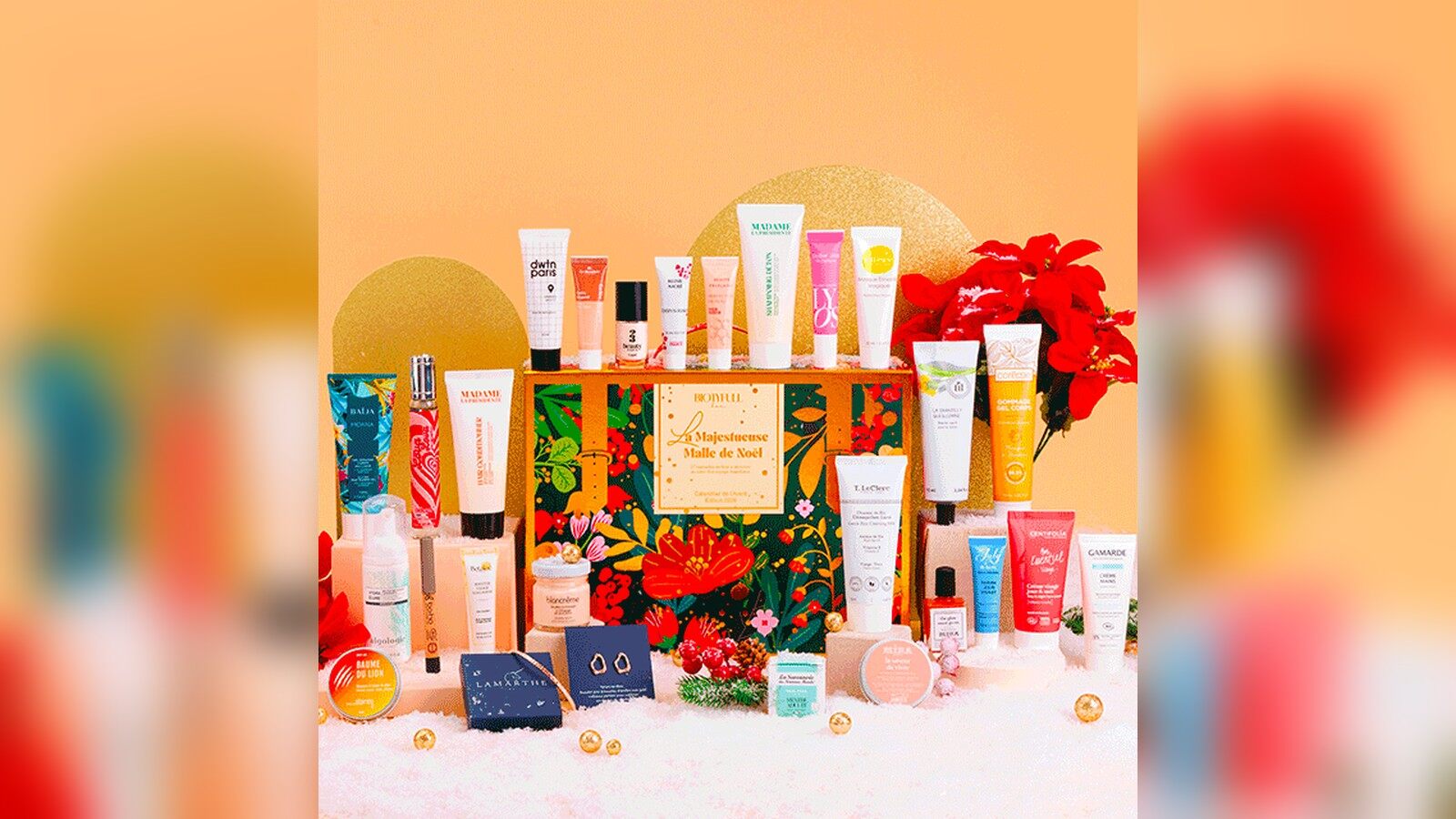 La Majestueuse Malle de Noël Biotyfull Box entourée de ses produits de beauté sur un décor orangé et de la neige artificielle