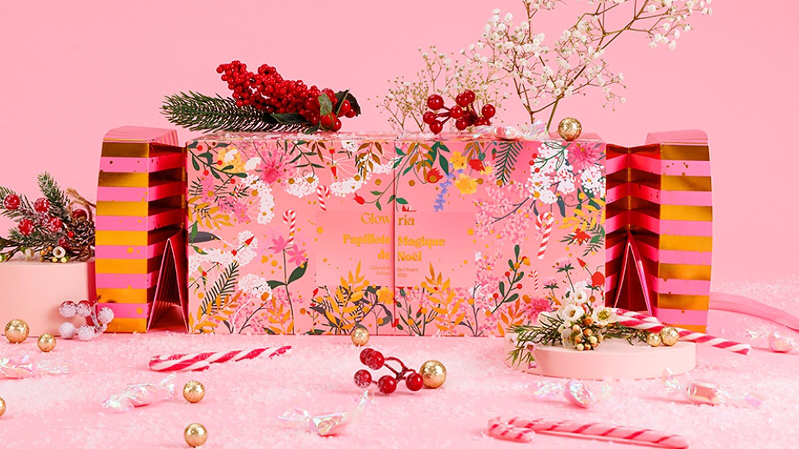 Le calendrier de l’Avent Glowria 2026 en forme de papillote géante rose et dorée, décor fleuri, sur un fond rose enneigé