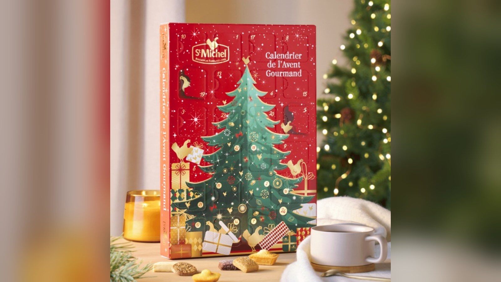 Le calendrier de l’Avent gourmand St Michel 2026, un coffret rouge à sapin vert, posé devant un sapin illuminé avec des biscuits et une tasse