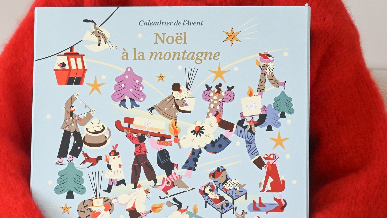 Une femme en pull rouge tient le calendrier de l’Avent My Jolie Candle 2026 illustré Noël à la montagne
