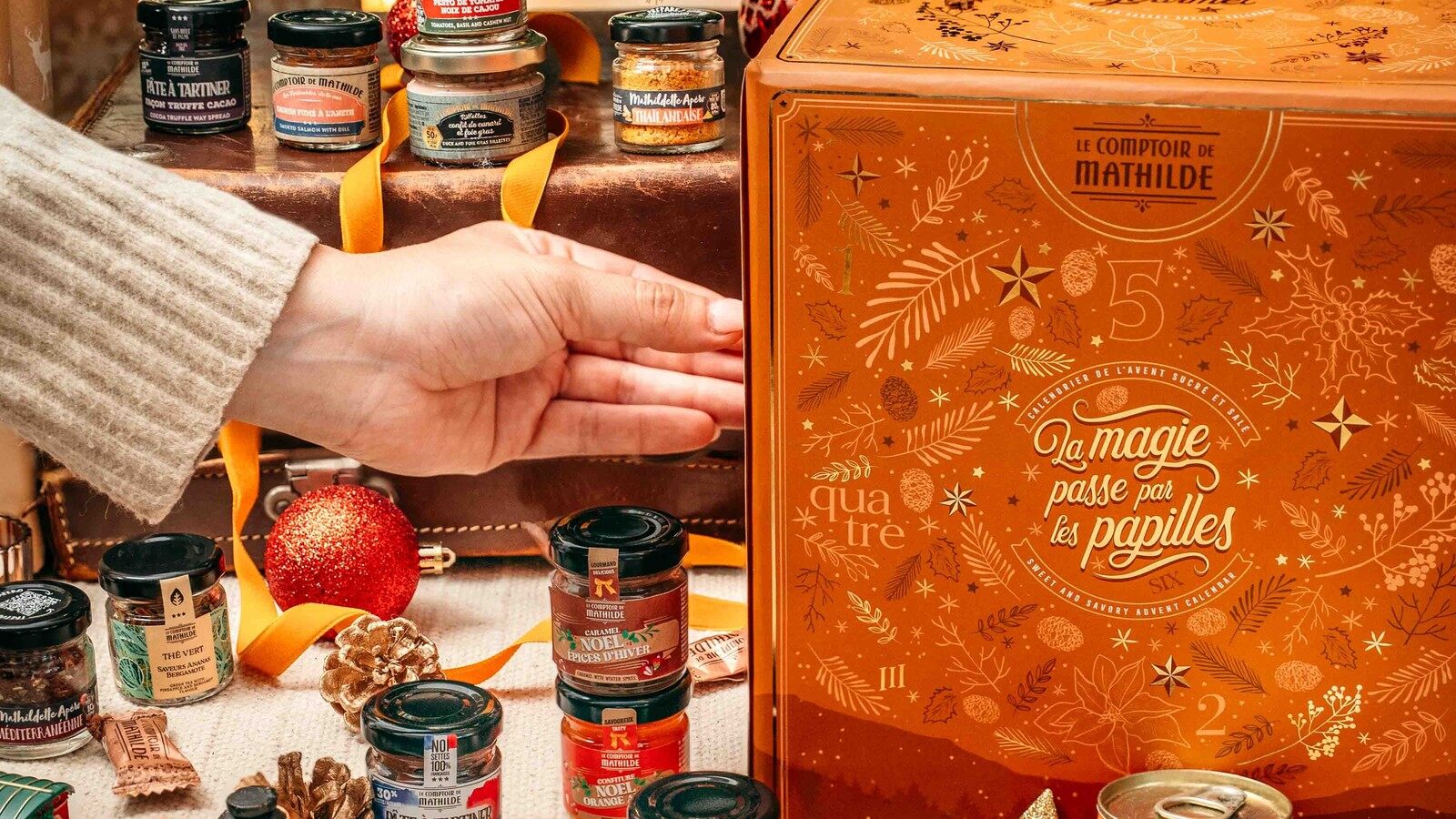 Une main pose des pots de tartinables devant le calendrier Gourmet du Comptoir de Mathilde, entouré de décorations de Noël
