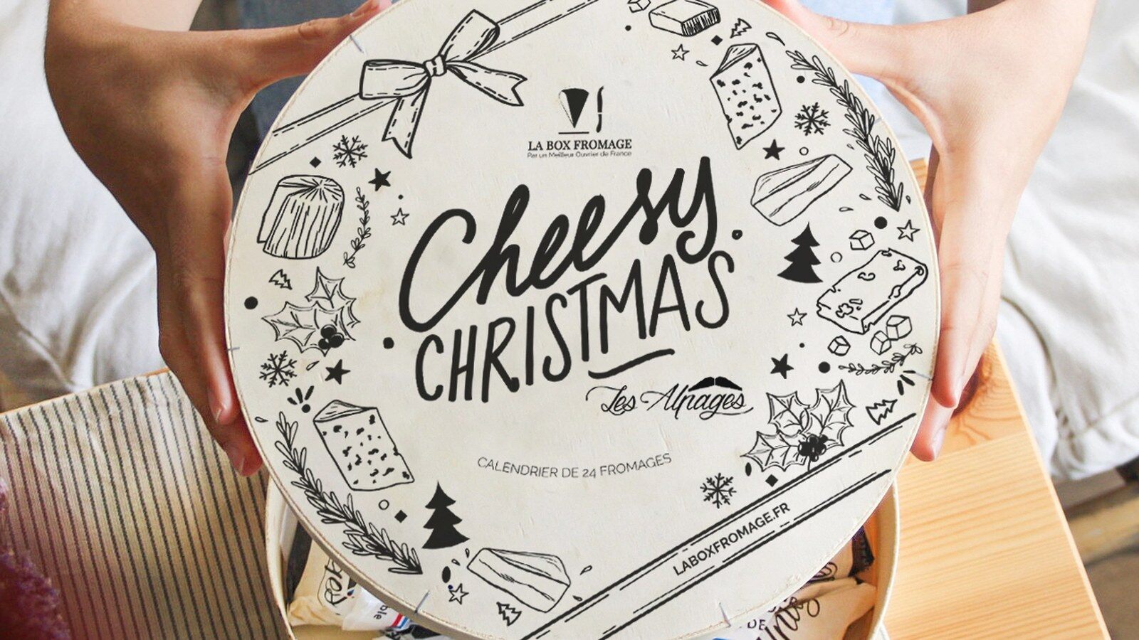 Deux mains ouvrent la boîte ronde en bois illustrée du calendrier de fromages Cheesy Christmas