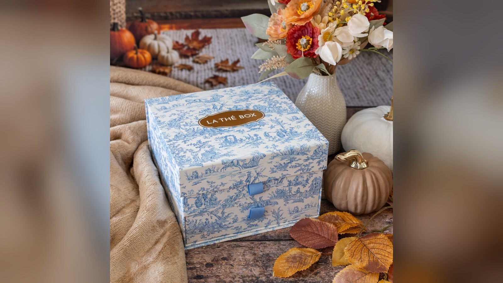 Le coffret Maison de Famille de La Thé Box, une boîte à tiroirs en toile de Jouy bleue, posée parmi des citrouilles, des feuilles d’automne et un bouquet
