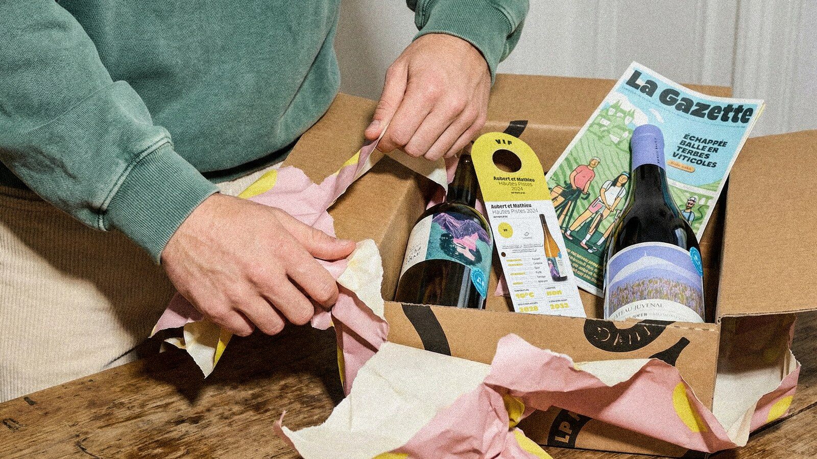 Deux mains déballent une box Le Petit Ballon avec deux bouteilles de vin et La Gazette