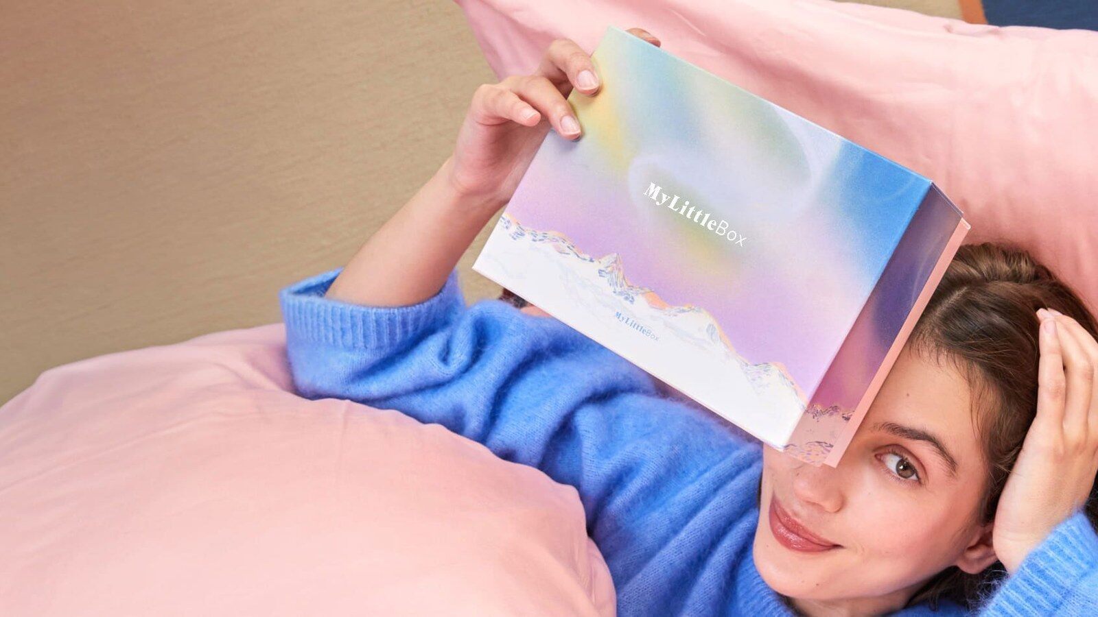 Une femme allongée sur des coussins roses tient une box My Little Box devant son visage